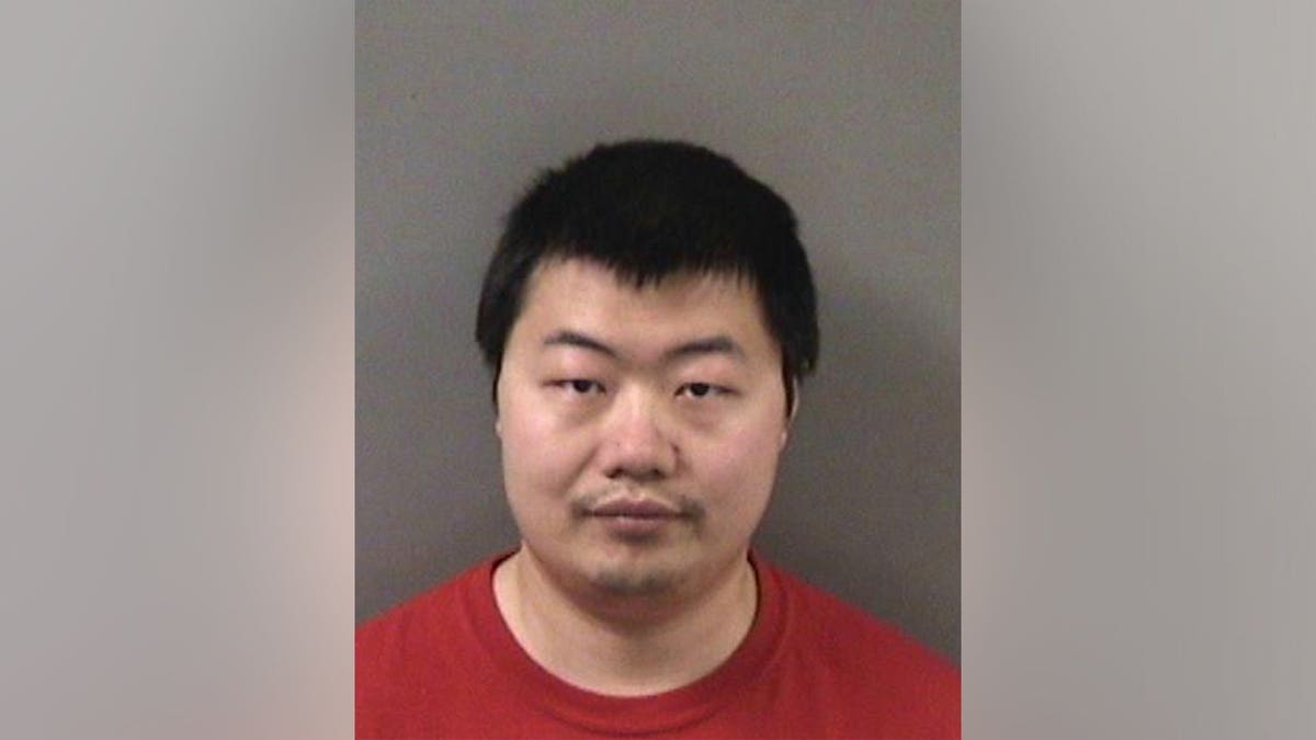 Booking photo of a man with short black hair wearing a red shirt, standing in front of a gray wall.