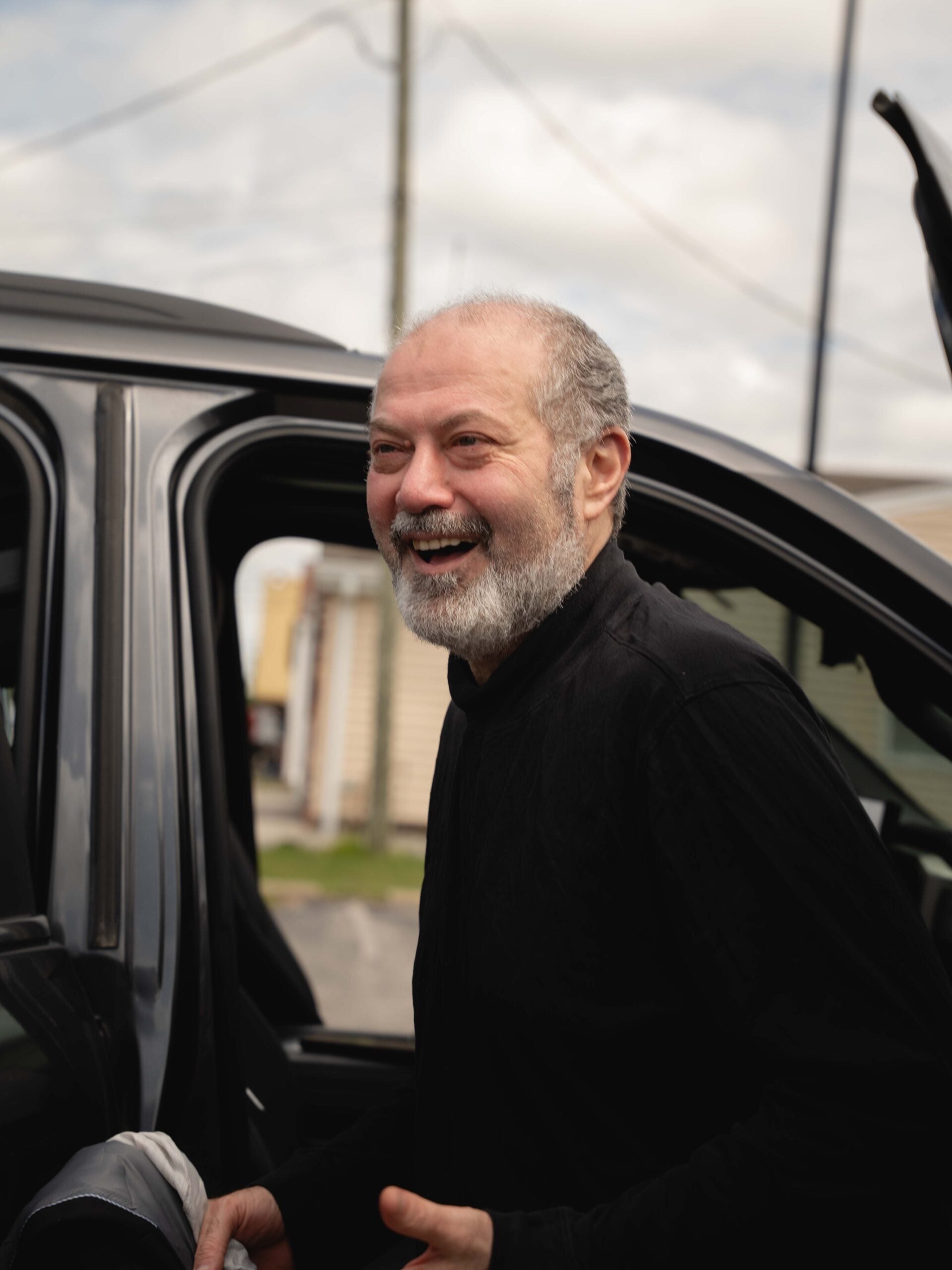A man with a gray beard and black shirt stands next to an open car door, smiling outdoors on a cloudy day.