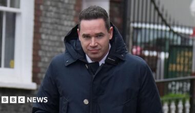 A man with short black hair wearing a large dark blue coat. He is looking into and walking towards the camera