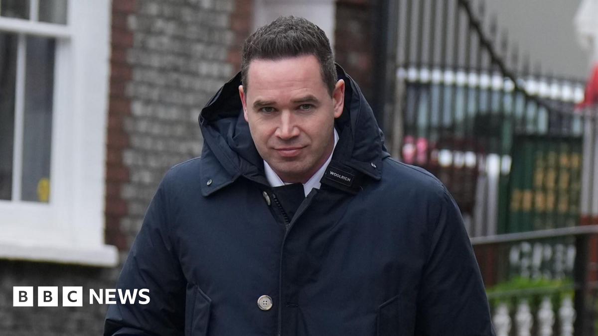 A man with short black hair wearing a large dark blue coat. He is looking into and walking towards the camera