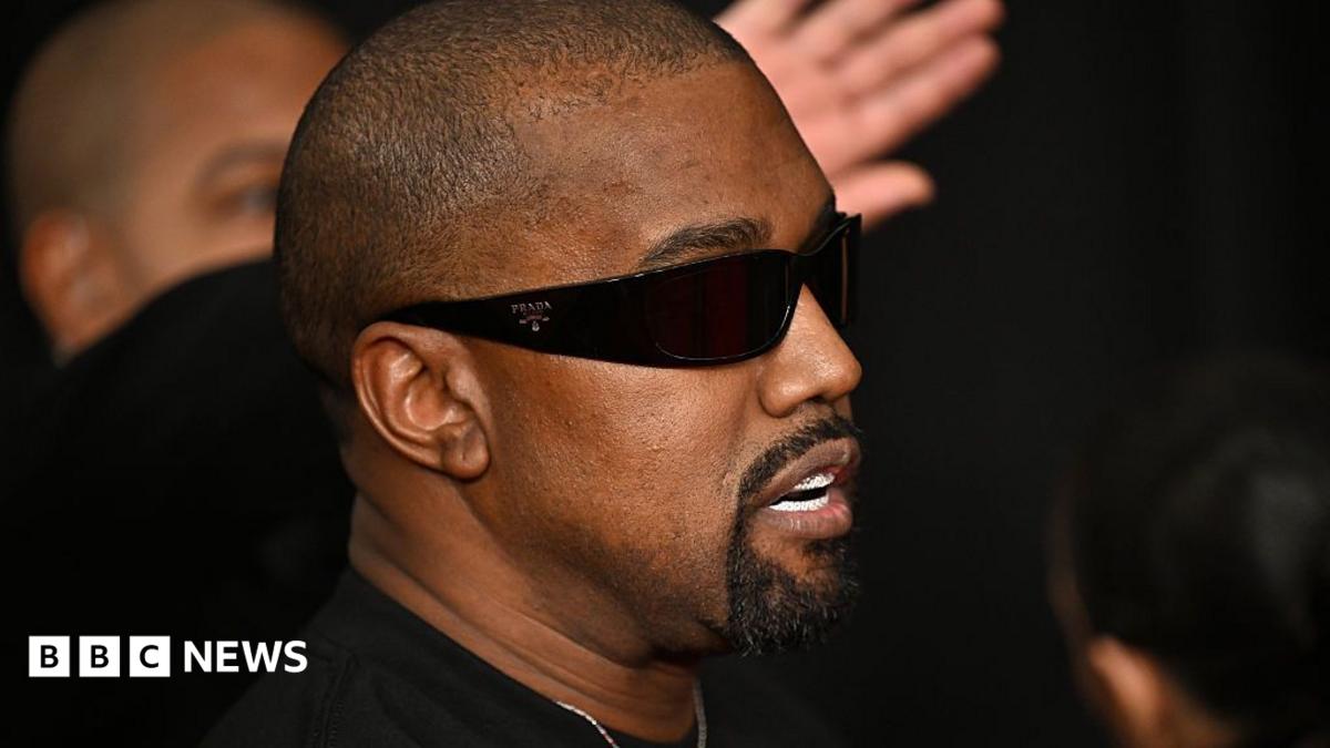 Kanye West in 2025 at the Grammy Awards. He has a shaved heard and a black goatee beard. He wears dark sunglasses and a black T-shirt