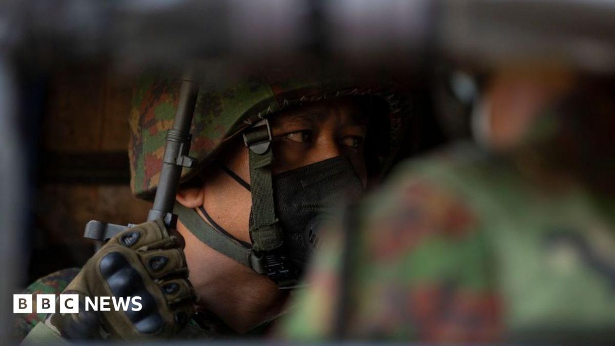 A soldier wearing a helmet and face mask looks to the side, his gloved hand holding the barrel of a rifle, his face only partially visible over the shoulder of another soldier.