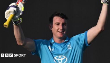 Harry Brook in a blue England kit. He is smiling with raised hands, holding his bat in one glove