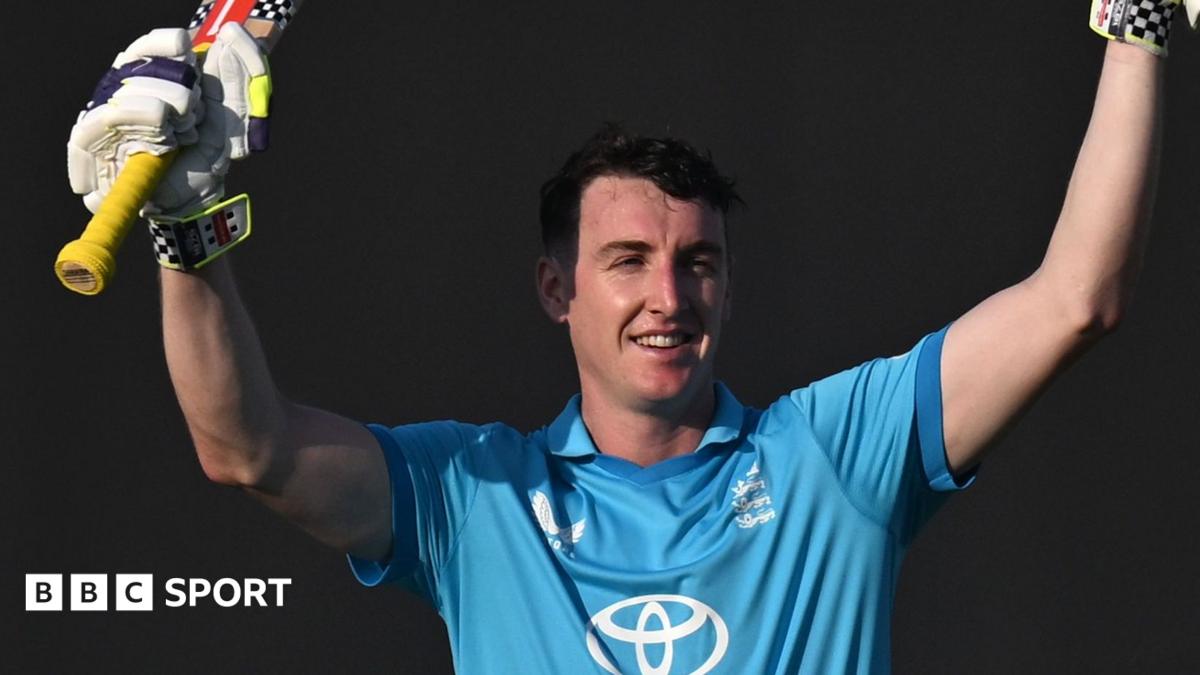 Harry Brook in a blue England kit. He is smiling with raised hands, holding his bat in one glove