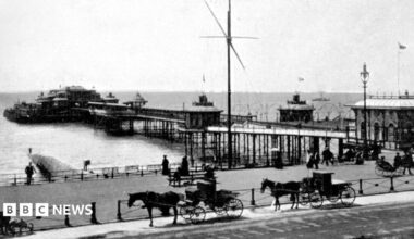 Large waves crashing over a black and white striped lighthouse at the end of a pier.