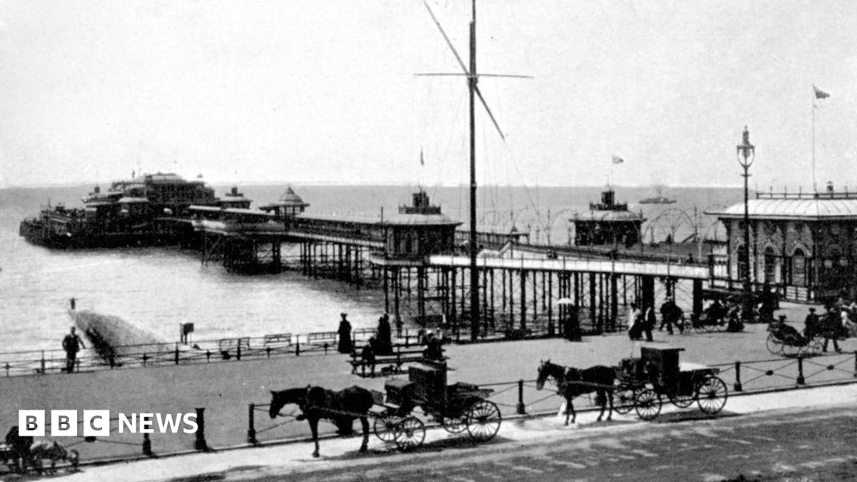 Large waves crashing over a black and white striped lighthouse at the end of a pier.