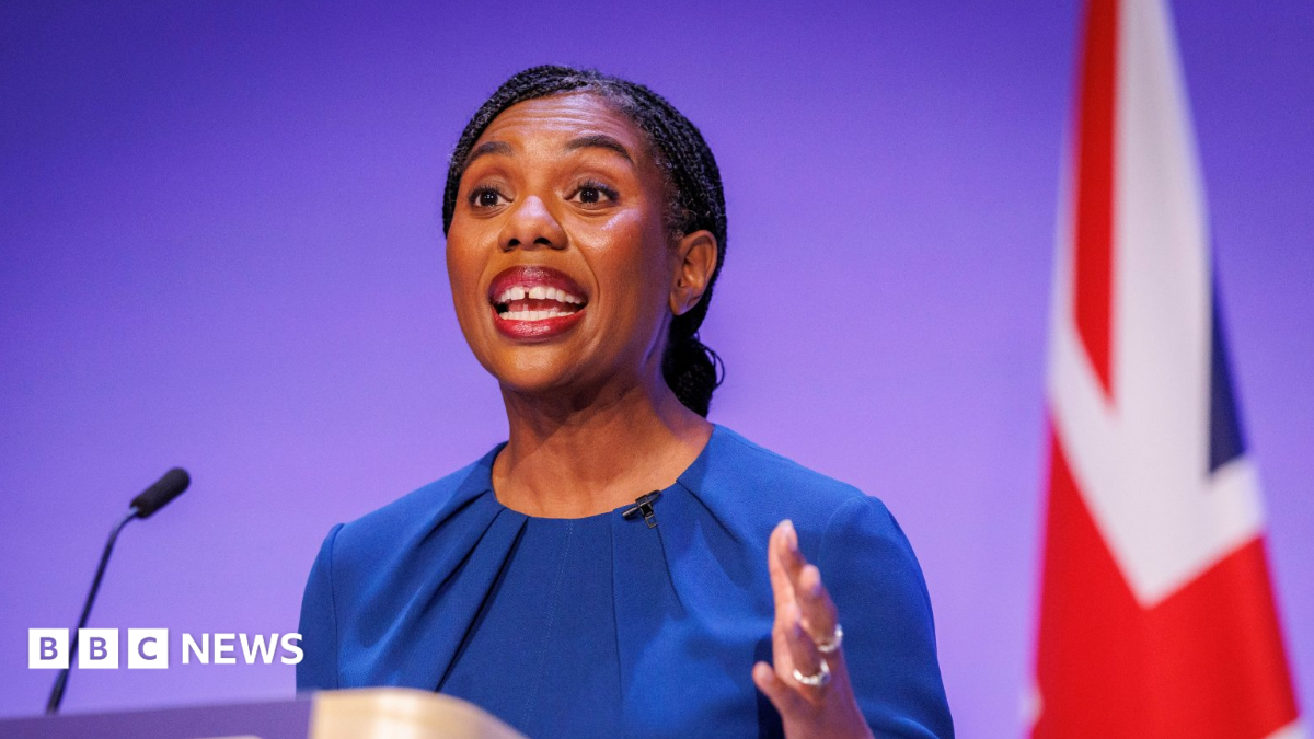 Hamish Badenoch and Kemi Badenoch are surrounded by Tory members, some of whom are holding tiny miniature Union flags. Hamish and Kemi Badenoch are both waving. Hamish is wearing a dark-coloured suit, white shirt and blue tie, while Kemi is wearing a blue dress.