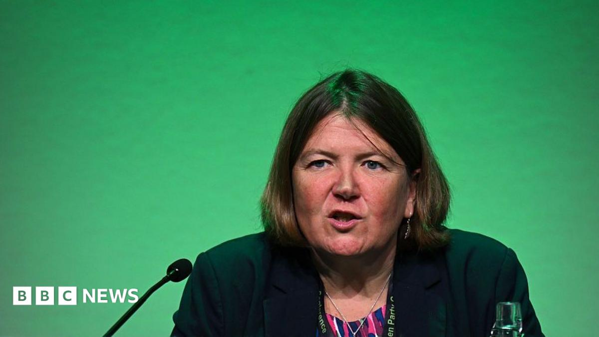 Ellie Chows in front a green background. She is speaking into a microphone, there is also a glass bottle of water on the desk she sits behind.