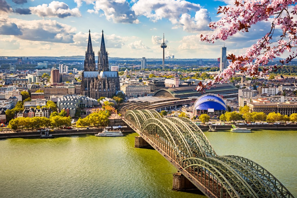 Aerial view of Cologne, Germany, showcasing the Rhine River, the Hohenzollern Bridge, and the Cologne Cathedral under a partly cloudy sky, with blooming cherry tree branches in the foreground.