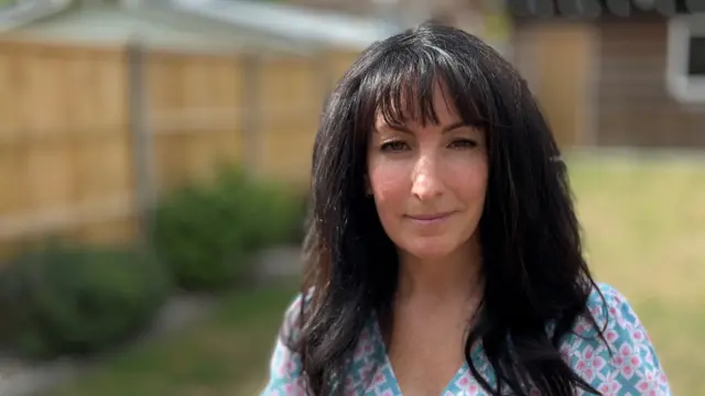A dark-haired woman wearing a floral-patterned shirt is sitting in the garden