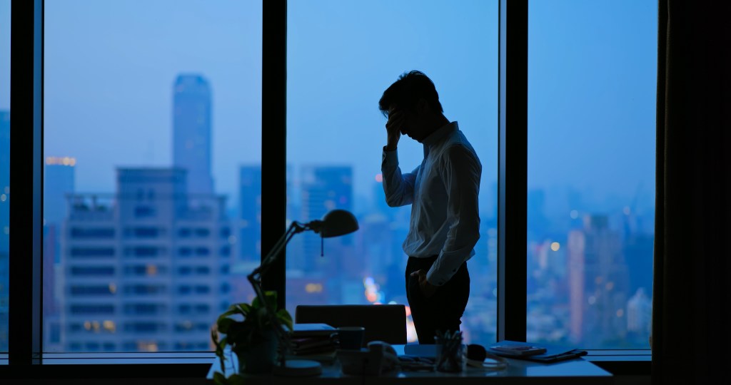 A frustrated businessman stands in a high-rise office at night, holding his head in his hand while looking out the window at the city skyline.