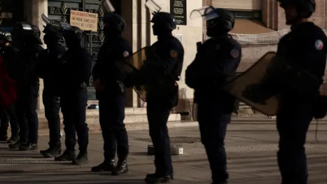 Eight black-clad riot police with helmets and shields stand in a line, with a protest sign in the background.