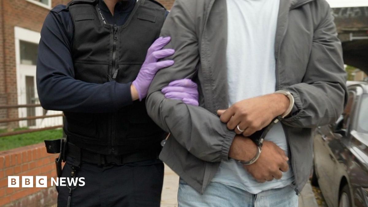 An officer wearing purple plastic gloves holds the arm of a man who is wearing handcuffs. they are walking along a pavement. The man is wearing a grey jacket, white t-shirt, light blue jeans and a ring on his left hand ring finger.