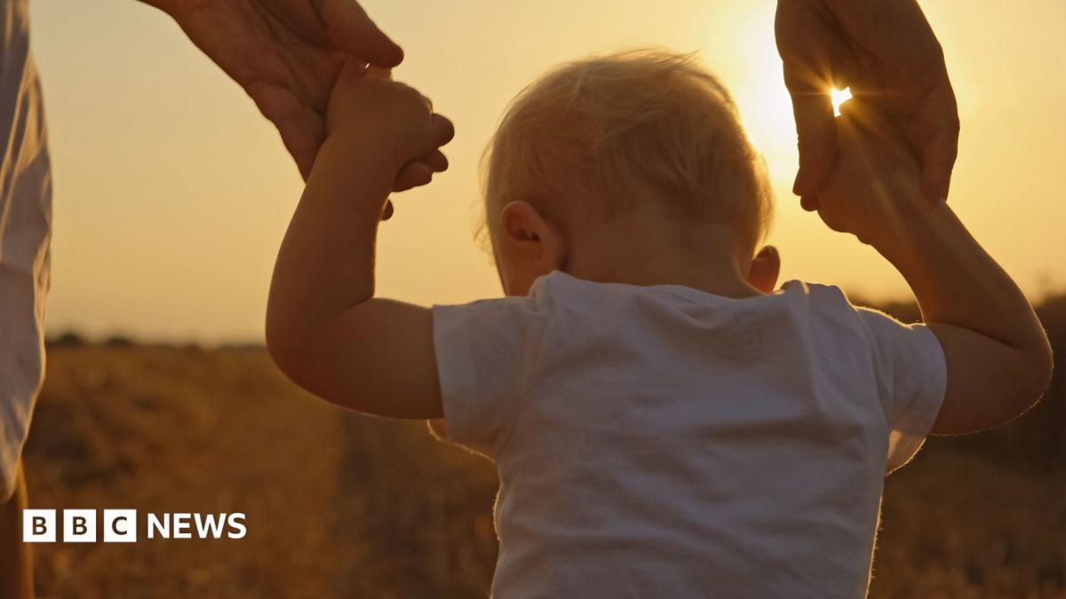 A small child, with blond hair and wearing a white T-shirt, being supported by two adult hands while outdoors in warm evening light.