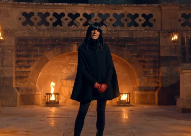 Claudia in a black cloak in front of two small fire pits and a brick wall