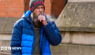 A man in his 30s, with a large beard, grey woollen hat and blue bubble coat, stands outside a stone building holding a black vape to his mouth.