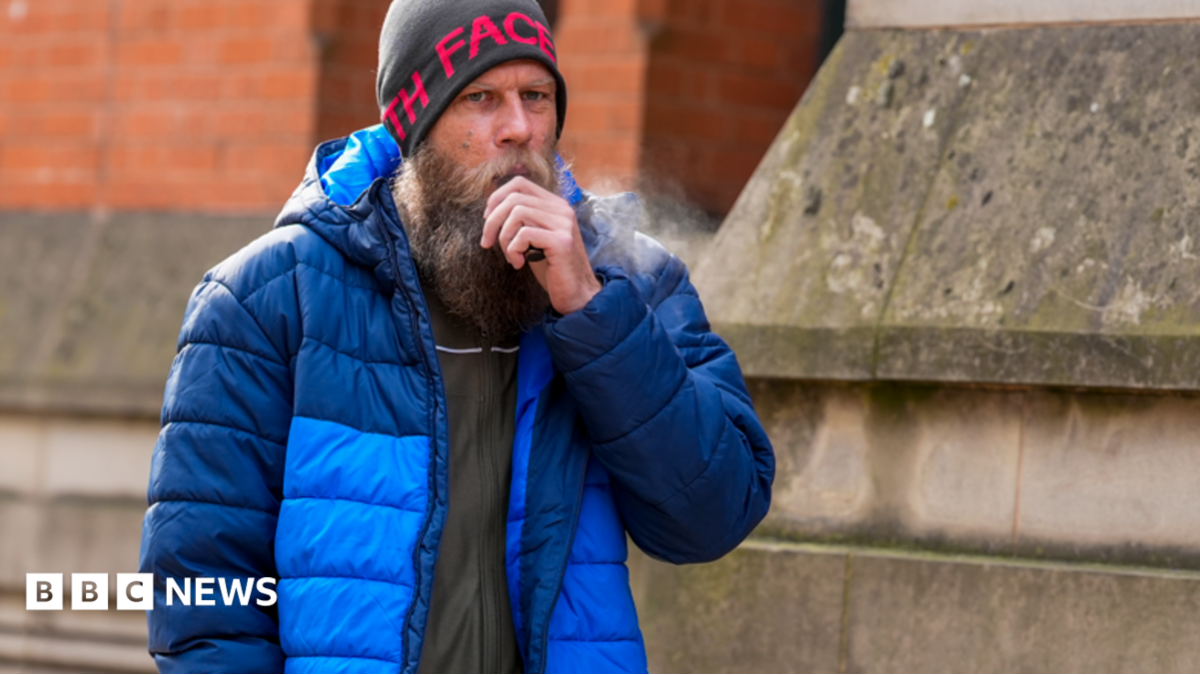 A man in his 30s, with a large beard, grey woollen hat and blue bubble coat, stands outside a stone building holding a black vape to his mouth.
