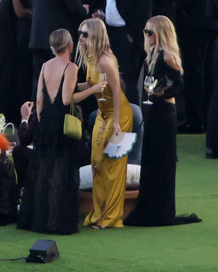 Rachel Zoe stands on the right wearing a black dress and sunglasses, holding a drink and a small clutch at an outdoor event.