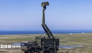 A green military vehicle sits in a field - on top of the vehicle is a tall green defence system that reaches into the sky. The ocean is visible in the distance.