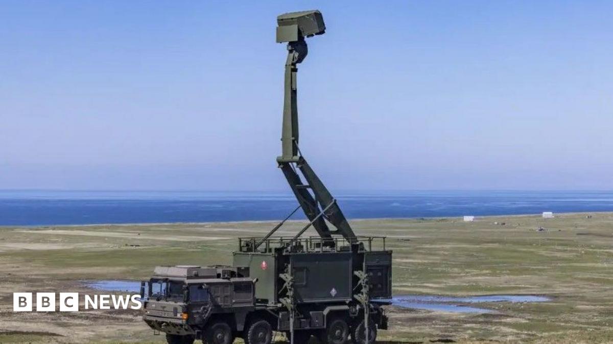 A green military vehicle sits in a field - on top of the vehicle is a tall green defence system that reaches into the sky. The ocean is visible in the distance.