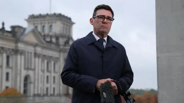Andy Burnham stands behind a microphone while wearing a suit and dark jacket.