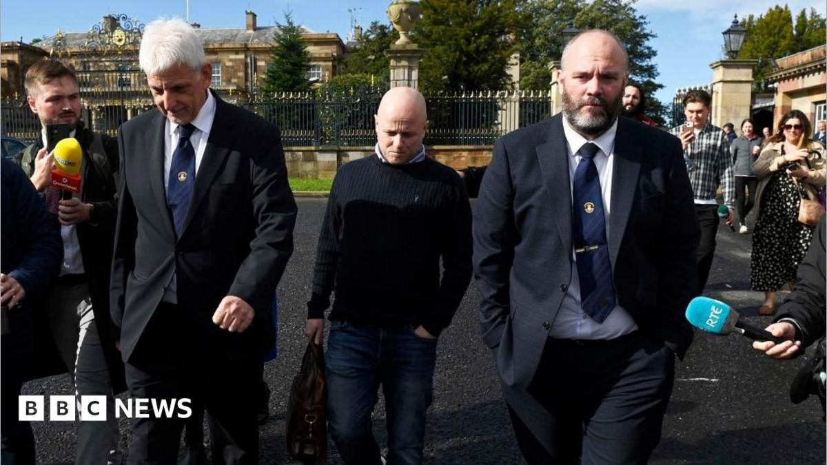 Three men walk side by side as members of the media point microphones in their direction. Two of the men are wearing dark suits, white shirts and navy ties. The other man is wearing a navy jumper and blue jeans.