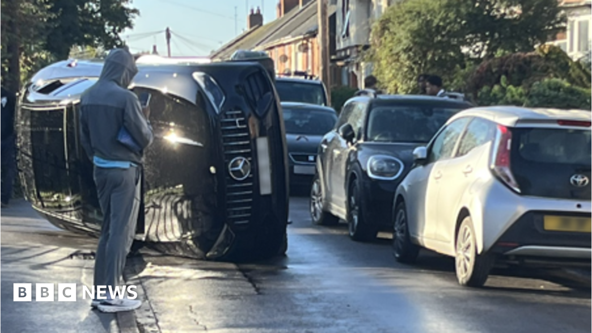 A black Mercedes is on its side in the middle of the road. There is a row of parked cars on the right hand side and a person in a grey sports tracksuit is standing in front of the car and is speaking into his phone.