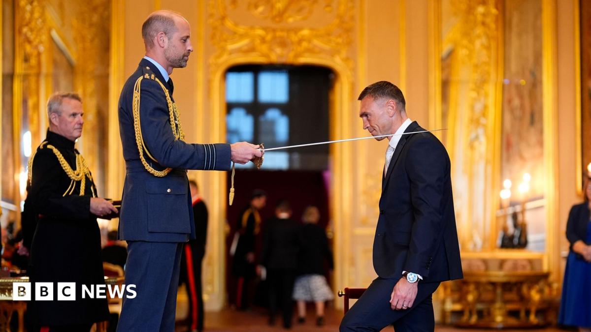 The Prince of Wales knights Sir Kevin Sinfield with a sword at an investiture in a grand, gold room at Windsor Castle.