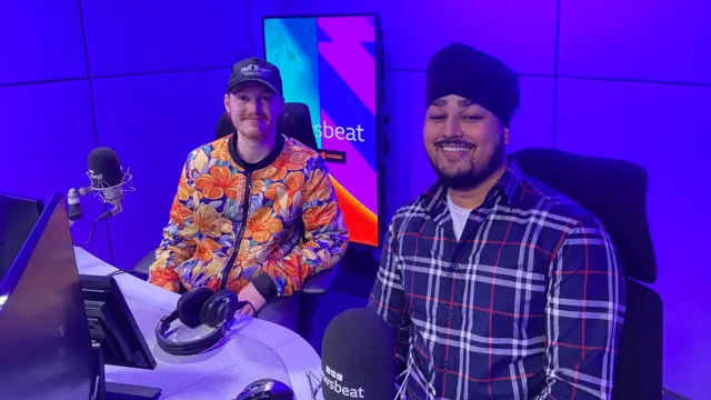 Two men sit in the BBC Newsbeat studio with microphones in front of them and blue lighting around them