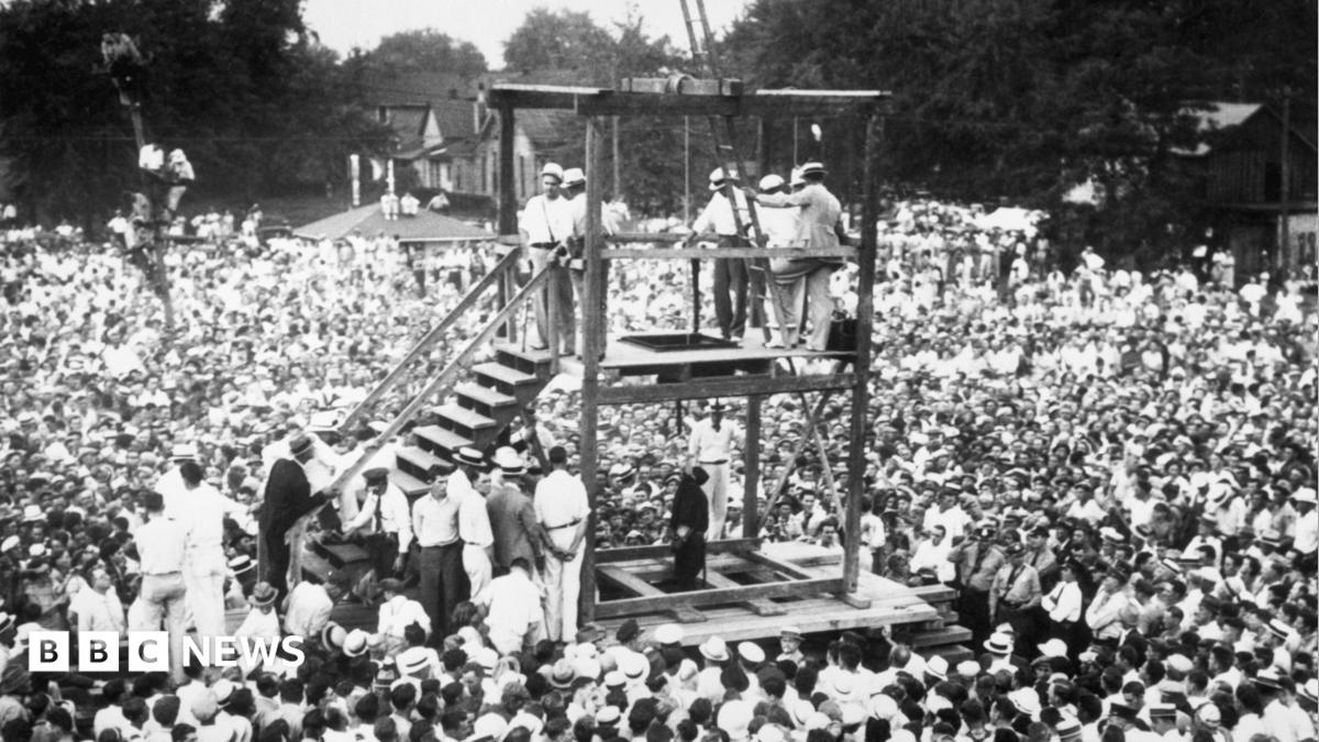 Men stand on wooden gallows, looking through a trap door at a black-clad figure hanging from a rope, as throngs of onlookers surround them