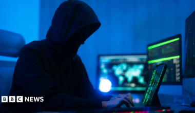 A person wearing a hooded coat is typing on a computer. They have multiple screens open and appear to be doing something covert. The room is illuminated with a dark blue light.