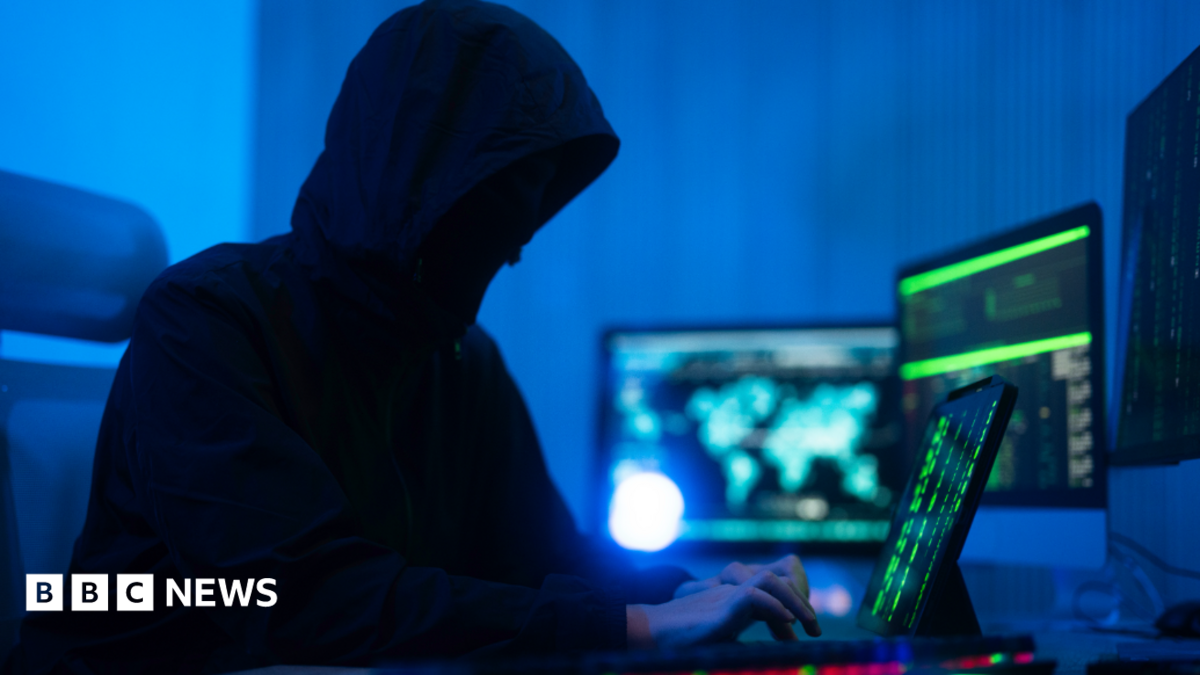 A person wearing a hooded coat is typing on a computer. They have multiple screens open and appear to be doing something covert. The room is illuminated with a dark blue light.