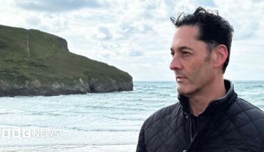 Simon Andriesz wearing a dark quilted jacket stands on a beach beside the sea. The photograph is taken in profile, with Andriesz looking toward the horizon. In the background, waves roll toward the shore and a grassy cliff extends into the water beneath a cloudy sky. The scene is brightly lit by daylight, with the coastline and ocean filling much of the frame.