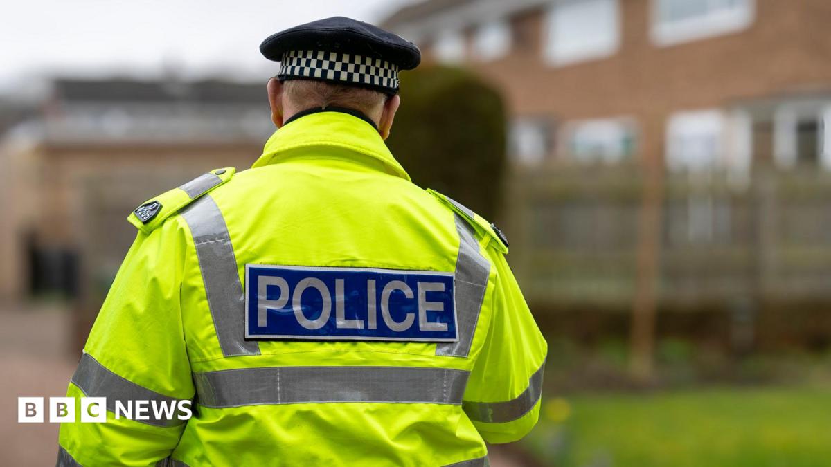 A police officer is walking away from the camera. He wears a yellow high-vis coat, which has silver reflective panels down the sides and a rectangular logo along the centre which reads 'POLICE' on a blue background. He also wears a black cap with a black and white check panel around the head. The background is blurred but shows a housing estate.