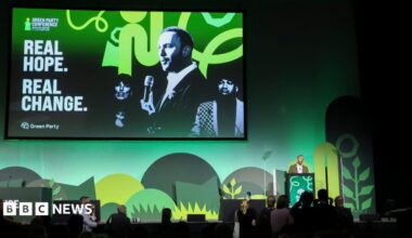 A stage with green shapes on a green background and a large screen depicting two men and a woman with the message "real hope real change" to the left of them. A man in an olive suit stands at a lectern in front of it.