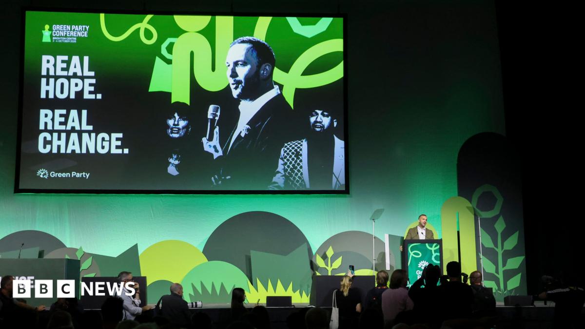 A stage with green shapes on a green background and a large screen depicting two men and a woman with the message "real hope real change" to the left of them. A man in an olive suit stands at a lectern in front of it.