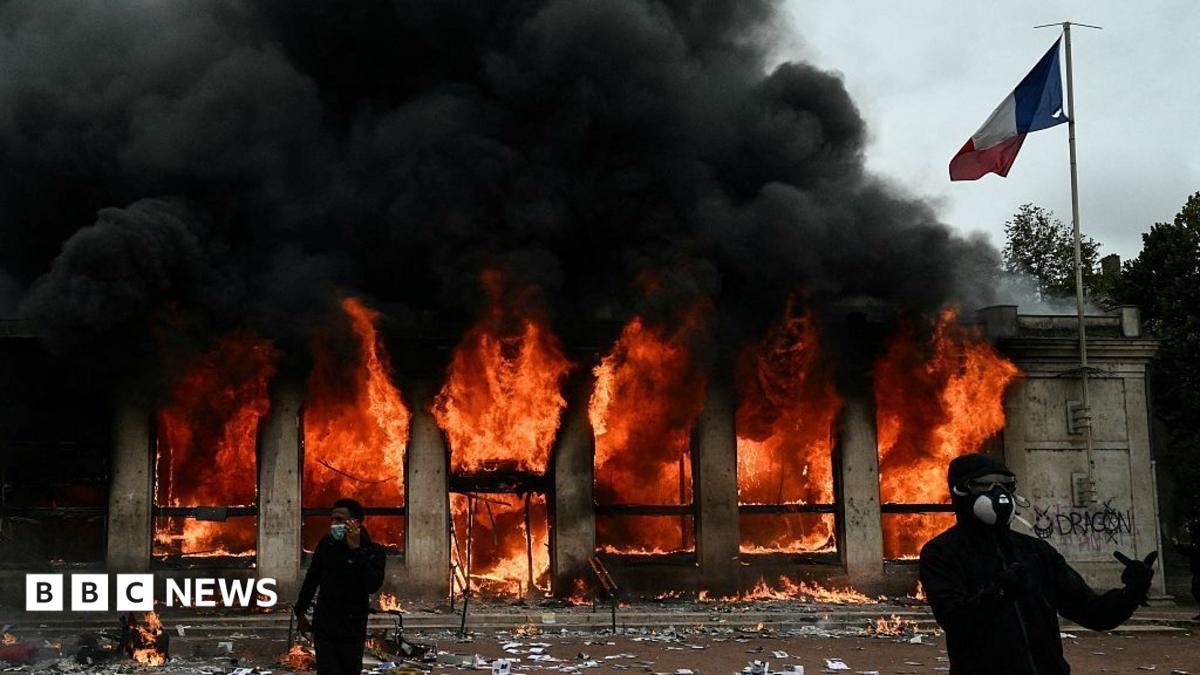 Two demonstrators, dressed all in black and in face coverings, stand outside the Lyon tourist office which is on fire behind them. A French flag is attached to the building.