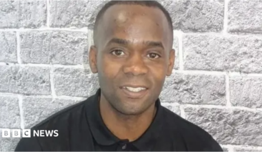 Portrait of Anton Green wearing a black long-sleeved polo shirt, in front of a light grey brick-pattern wall. He faces the camera, with his head and shoulders visible in the frame. He is smiling.