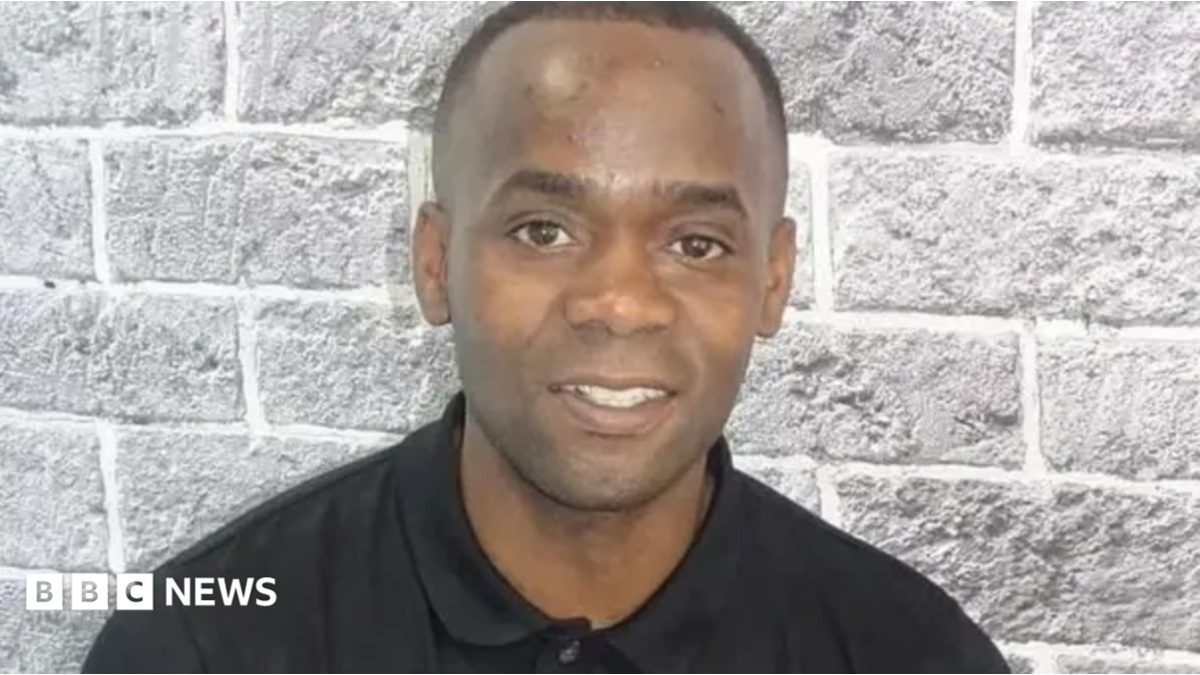 Portrait of Anton Green wearing a black long-sleeved polo shirt, in front of a light grey brick-pattern wall. He faces the camera, with his head and shoulders visible in the frame. He is smiling.
