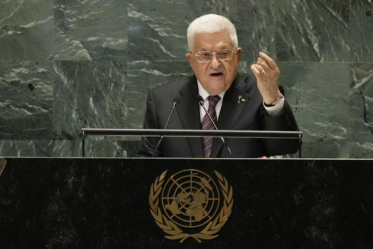 A white-haired man in a suit speaks from a podium.