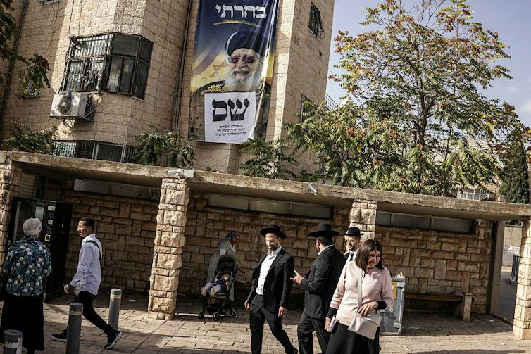 A few men walk through a plaza in front of a building with a large poster of a man with a white beard and dark glasses.