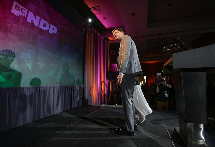 A tall man in a grey suit walks off a stage.