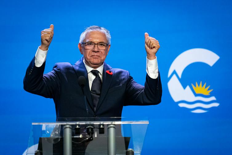 A man with grey hair awkwardly raises his fists in front of a blue Conservative party banner.