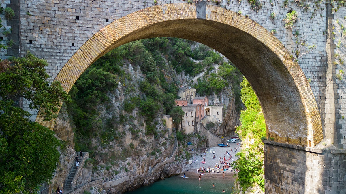 Idyllic view of Fiordo di Furore beach and cove in southern Italy, framed by an old arched bridge, with colorful buildings and swimmers below.