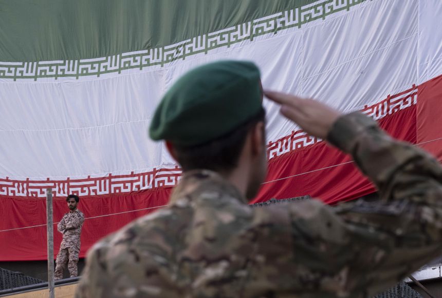 An Islamic Revolutionary Guard Corps serviceman salutes in front of an Iranian national flag.