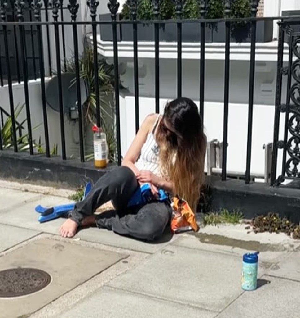 Mahrah Al Nahyan sits on the sidewalk leaning against a black metal fence, with snack bags and a bottle nearby.