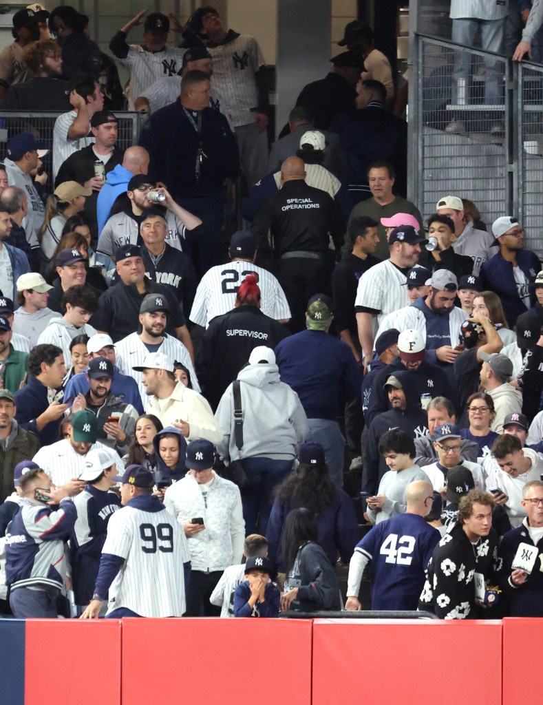 A crowded section of Yankee Stadium stands featuring security personnel and a fan in a white jersey being escorted out after interfering with a ball during a game.