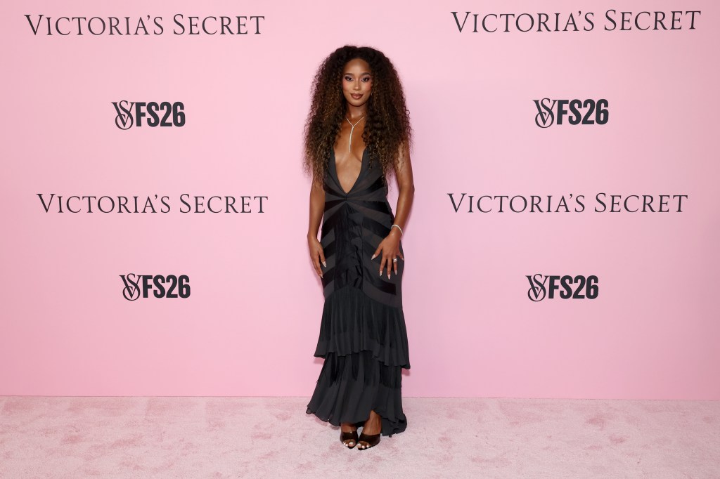Mia Calabrese poses on the pink carpet at the Victoria's Secret Fashion Show, wearing a dark plunging gown and layered necklaces.