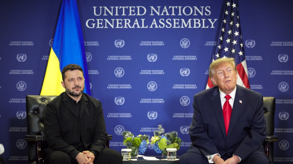 US President Donald Trump and Ukraine President Volodymyr Zelensky are both sitting in black chairs. They are both looking to slightly to their left sides toward something outside of the photo. In the background is one Ukraine, one US flag, and a blue press conference wall that reads "UNITED NATIONS GENERAL ASSEMBLY" in white letters.